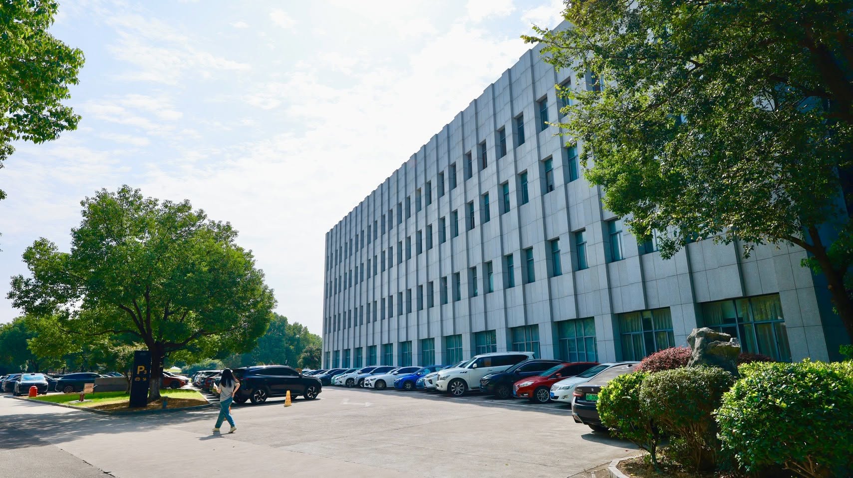 Nhà máy sản xuất pin lưu trữ APESS: Ấn tượng từ khuôn viên bên ngoàiẤn tượng đầu tiên của chuyến tham quan là khối nhà có mặt đứng trải dài, thiết kế nhiều tầng với những ô cửa kính được bố trí đều đặn. Tông xám sáng, đường nét vuông vức và các mảng kiến trúc lặp lại tạo cảm giác bề thế, chỉn chu.Phía trước là khoảng sân rộng, khu vực đỗ xe và những hàng cây lớn. Cây xanh được bố trí xen kẽ cùng thảm cỏ, bụi cây và các khoảng cảnh quan, giúp tổng thể khuôn viên thoáng đãng hơn.Sự kết hợp giữa công trình quy mô lớn và không gian xanh tạo nên diện mạo chuyên nghiệp cho điểm đến. Đây cũng là khung cảnh mở đầu hành trình tìm hiểu sâu hơn về hoạt động sản xuất pin lưu trữ APESS.